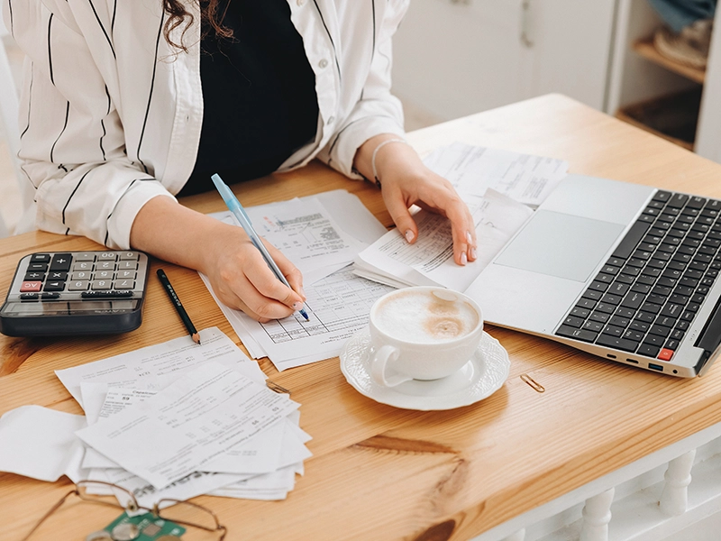 A person sits at a wooden desk with bills, a calculator, and coffee, writing on a document while referencing papers. An open laptop nearby suggests they are tackling business struggles or analyzing business profitability.