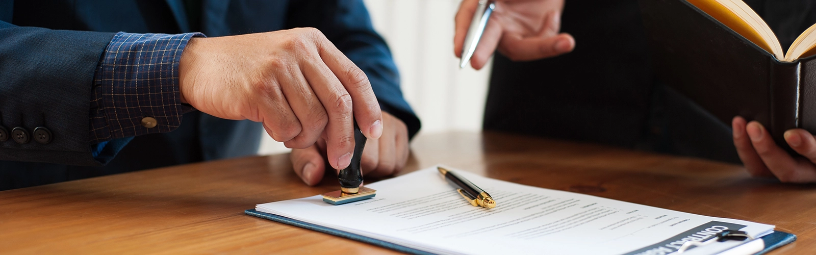 Two people are finalizing a contract at a wooden desk. One in a suit presses a stamp onto the document, while the other holds an open folder. The scene suggests a formal signing process, possibly for audit services or risk management assurance.